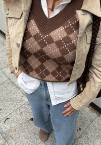 Person wearing beige suede jacket over brown argyle knit vest and white shirt, blue jeans, standing on stone pavement with leaves.