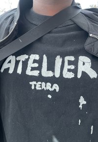 Melna kokvilnas T-krekls ar lielu, saplaisājušu baltu tekstu "ATELIER TERRA." Redzami pūki un traipi uz auduma virsmas.