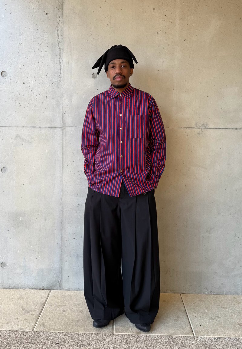 Uomo in piedi contro un muro di cemento che indossa un cappello nero con orecchie flosce, una maglietta a righe rosse e blu e pantaloni neri ampi.