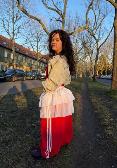 Mujer con cabello largo y rizado que lleva una chaqueta de mezclilla, una falda blanca con capas y pantalones rojos, de pie en un camino cubierto de césped con árboles sin hojas y coches cercanos.