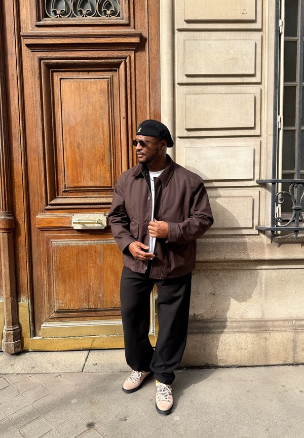 Homme portant un béret noir, des lunettes de soleil, une veste marron, un pantalon noir et des chaussures beige, debout devant une porte en bois et un bâtiment en pierre sous la lumière du soleil.