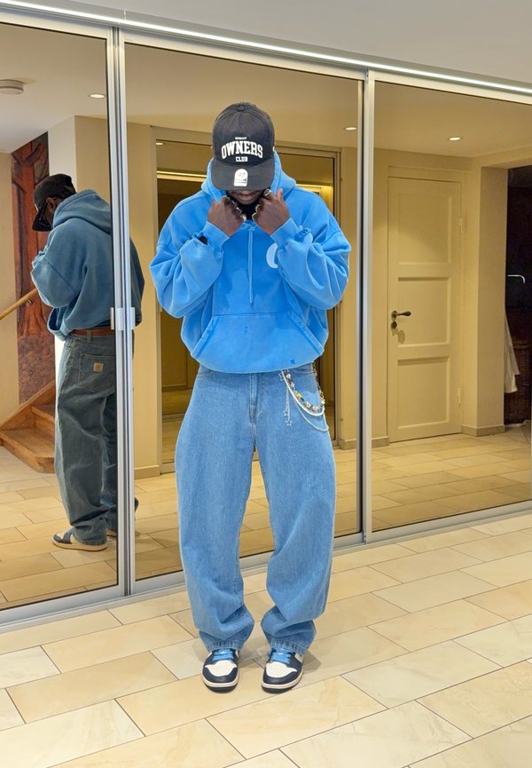 Homme portant un sweat à capuche bleu, un jean large, des baskets et une casquette noire debout à l'intérieur devant des portes coulissantes miroir.