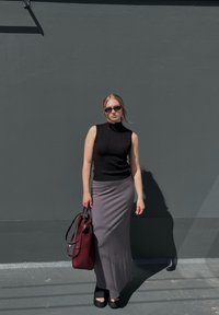 Črna rebrasta brezrokavnik, siva prilegajoča maxi krila, odprti čevlji na platformo, drži burgundsko torbo ob sivi steni.