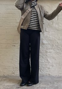 Jachetă de piele bej purtată peste un pulover tricotat cu dungi negre și albe, asortată cu pantaloni largi negri și balerini negri, pe fundalul unei pereți texturat.