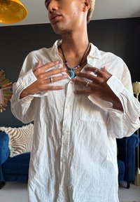 Personne portant une chemise texturée blanche ajustant un collier en forme de croissant bleu-vert, avec plusieurs bagues, devant un mur sombre et un canapé bleu en arrière-plan.