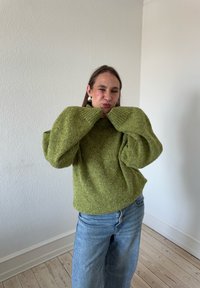 Túlméretezett zöld kötött pulóver magas nyakkal és széles ujjakra, világoskék farmernadrággal párosítva, egy sima fehér fal előtt.
