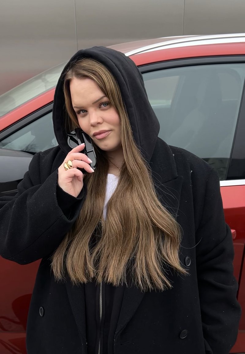 Jeune femme aux longs cheveux et sweat à capuche noir tenant un téléphone noir à son oreille, debout à côté d'une voiture rouge.