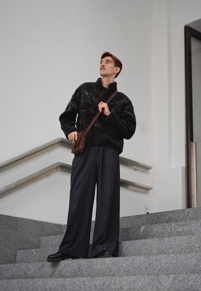 Sudadera negra de forro polar con cuello alto, pantalones oscuros de pierna ancha y bolso cruzado marrón. Fondo de escalera lisa con diseño minimalista.