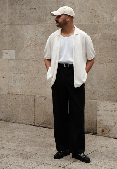 Camisa blanca de textura y manga corta sobre una camiseta blanca, combinada con pantalones negros de pierna ancha y mocasines negros. Una gorra beige completa el conjunto.