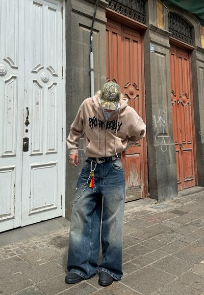 Jeans amplios de color azul desgastado con un cinturón negro con cadenas, combinados con una sudadera rosa clara y una gorra de camuflaje. Fondo de hormigón con puertas.