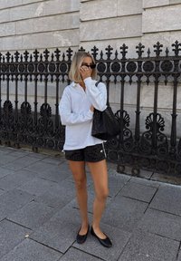 Hvit langermet polo skjorte, svarte stripete shorts, svarte ballerinasko og en strukturert svart tote-veske mot en steinvegg og jernrekkverk.