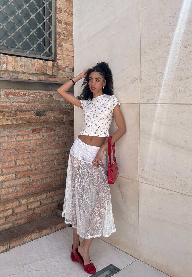 Femme portant un haut blanc à motifs floraux et une jupe en dentelle transparente, tenant une pochette rouge, debout contre des murs en briques et en pierre.