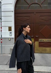 Šedý vlněný zkrácený kabát s širokými rukávy, nošený přes černý top. Vyznačuje se svou jemnou strukturou a jednoduchým designem, umístěným na pozadí dřevěných dveří.