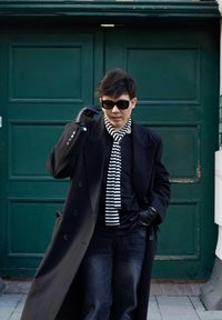 Manteau long noir à grands revers, gants noirs et écharpe rayée. Jean foncé, lunettes de soleil. Porte verte avec un design à panneaux en arrière-plan.