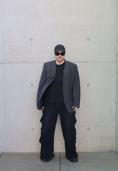Hombre con blazer gris oversize, pantalones cargo negros, gorra negra y gafas de sol oscuras, apoyado contra una pared de hormigón.