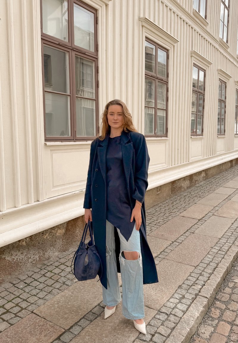 Mujer con un abrigo azul marino y jeans rotos de color azul claro sostiene un bolso azul marino, parada en una acera de adoquines junto a un edificio de color crema.