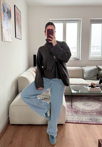 Suéter oversized gris oscuro con puños acanalados, jeans de pierna ancha azul claro y zapatillas turquesas con rayas blancas, posado en una habitación bien iluminada.