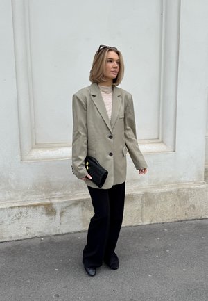 Blazer de gran tamaño gris claro, pantalones negros de campana y un bolso de mano negro texturizado. El conjunto incluye collares en capas y gafas de sol cuadradas.