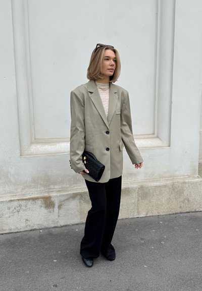 Blazer de gran tamaño gris claro, pantalones negros de campana y un bolso de mano negro texturizado. El conjunto incluye collares en capas y gafas de sol cuadradas.
