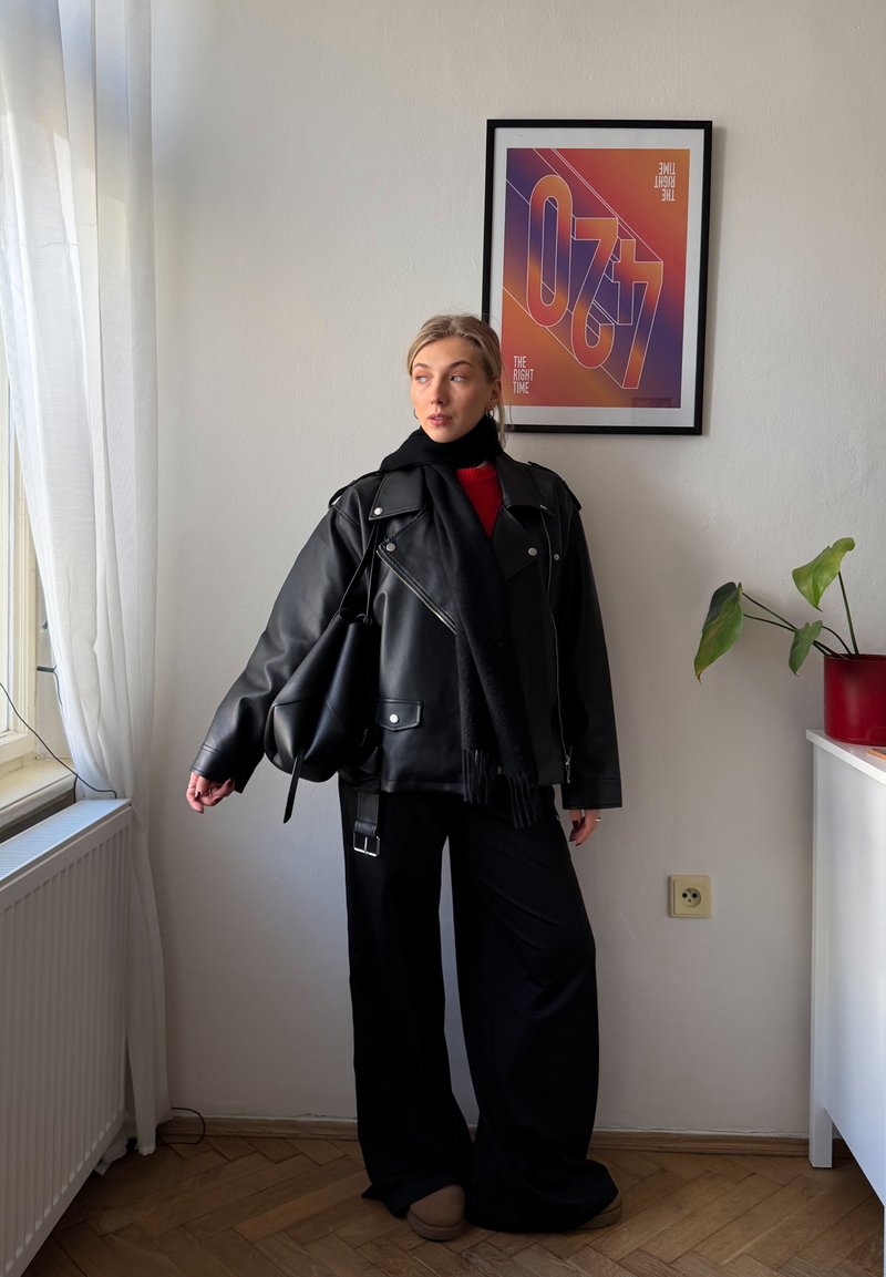 Jeune femme en veste en cuir noire et écharpe se tient à l’intérieur près d’une fenêtre avec une affiche colorée encadrée et une plante en pot en arrière-plan.
