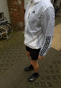 Veste légère blanche avec capuche, ornée de bandes noires sur les manches, associée à un short noir et des chaussures de sport noires sur une surface pavée.