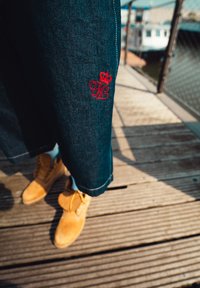 Tmavě modré džínové kalhoty s červeným vyšitým logem s písmeny "TJ" a korunou. Spárované s béžovými semišovými botami.