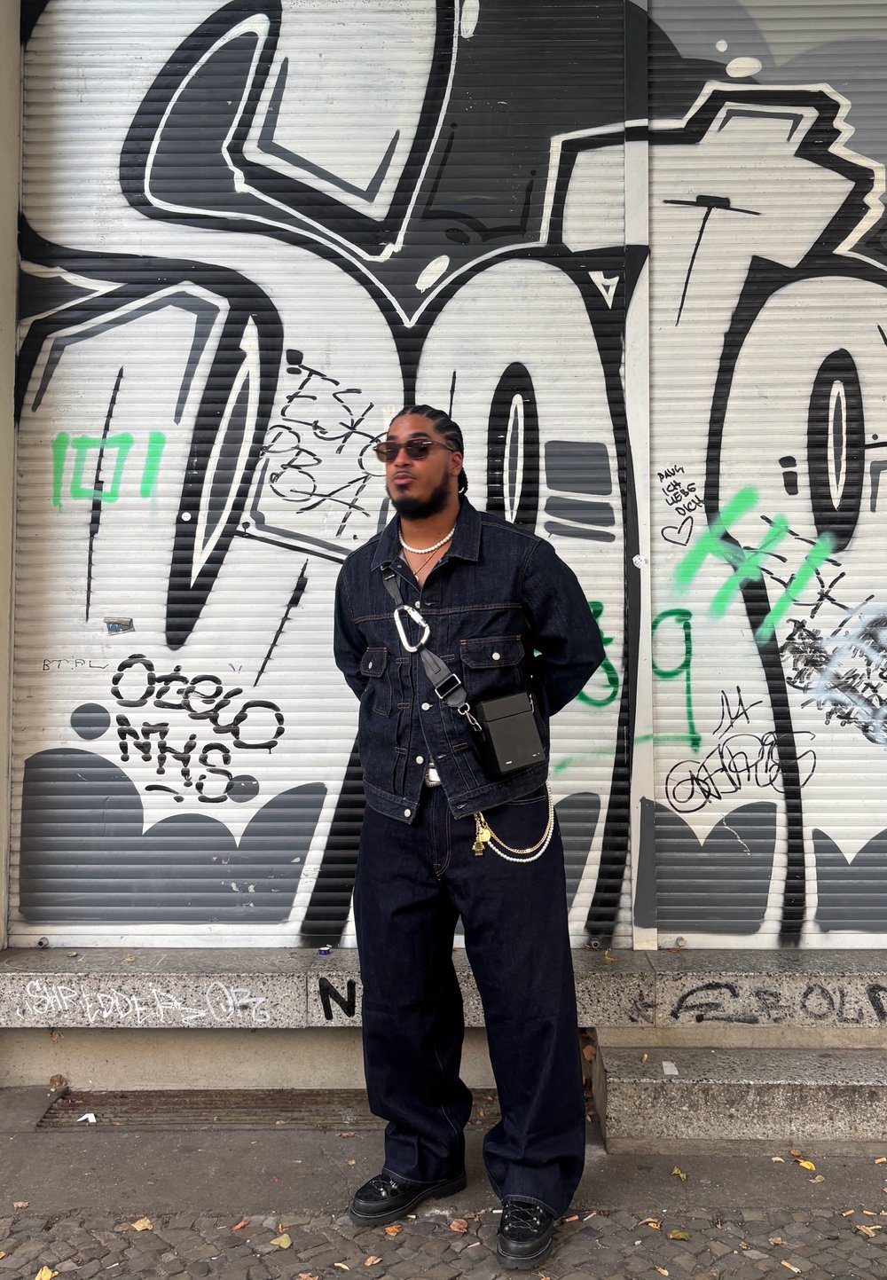Veste en denim foncé avec un jean large assorti. Sac bandoulière noir texturé, accents en chaîne argentée, portant des lunettes de soleil rondes noires. Fond de graffiti urbain.
