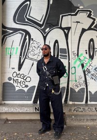Veste en denim foncé avec un jean large assorti. Sac bandoulière noir texturé, accents en chaîne argentée, portant des lunettes de soleil rondes noires. Fond de graffiti urbain.