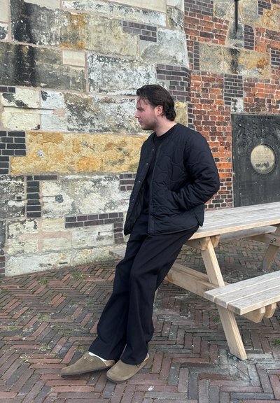 Chaqueta acolchada negra, pantalones negros y zapatillas marrones. Apoyado contra una pared de piedra con un banco de madera en un entorno de adoquines.