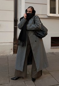 Lange grijze wollen jas met wijde mouwen, gecombineerd met een zwarte sjaal en puntige zwarte hakken. Een grote zwarte handtas met studs hangt over de schouder.