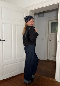 Czarna krótka kurtka, szerokie ciemnoniebieskie dżinsy z mankietami oraz wzorzysty czarny bandana. Wnętrze z białymi drzwiami.