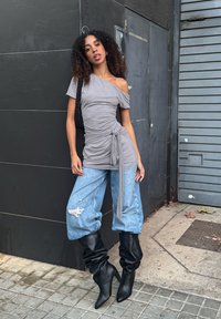 Sivi top s otkrivenim ramenima i skupljenim strukom, uparena s plavim širokim trapericama s poderanim detaljima. Nošeno s crnim čizmama do koljena.