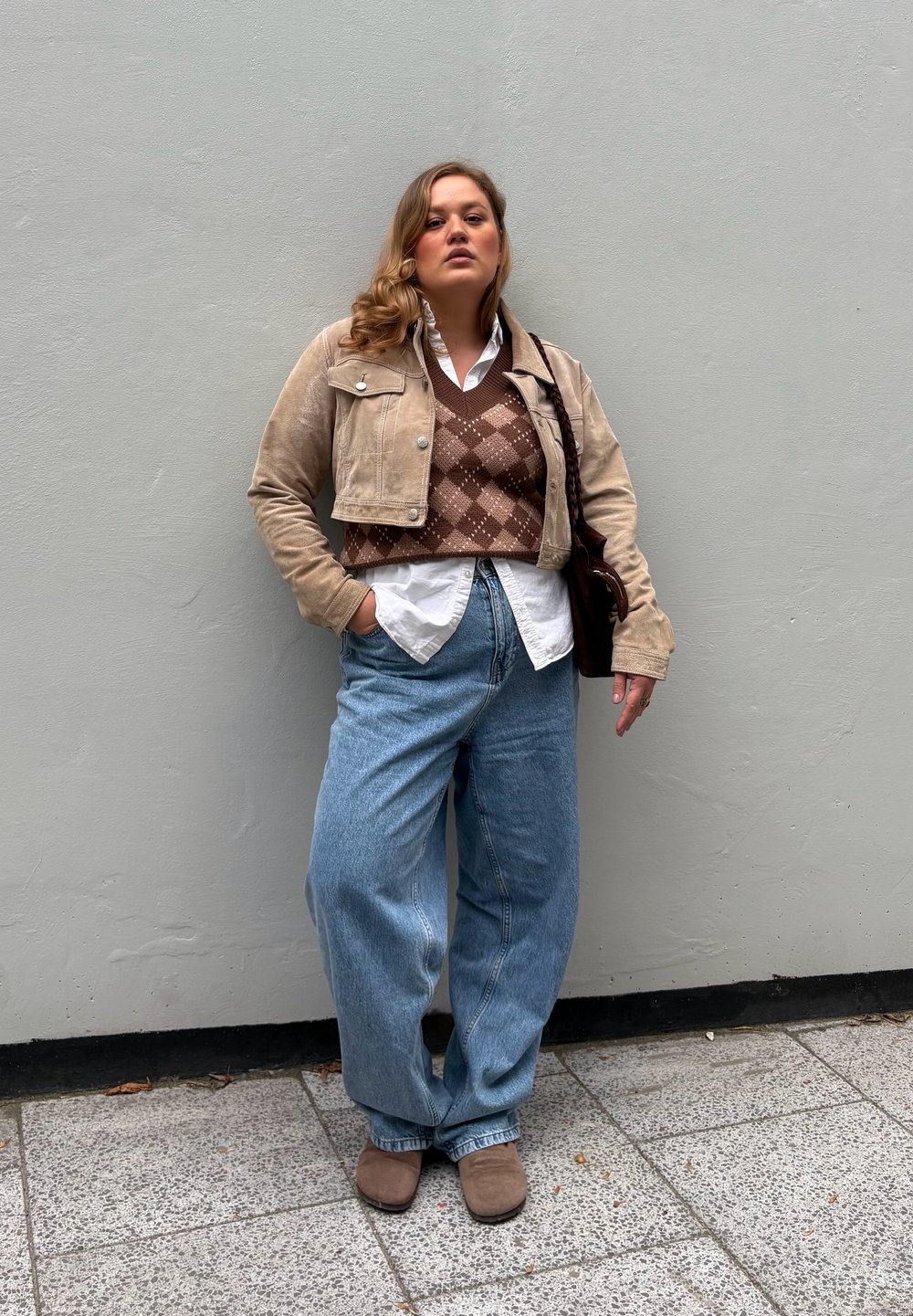 Woman standing against gray wall wearing beige cropped jacket, brown argyle sweater vest, white shirt, wide jeans, and brown boots with hand in pocket.