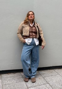 Woman standing against gray wall wearing beige cropped jacket, brown argyle sweater vest, white shirt, wide jeans, and brown boots with hand in pocket.