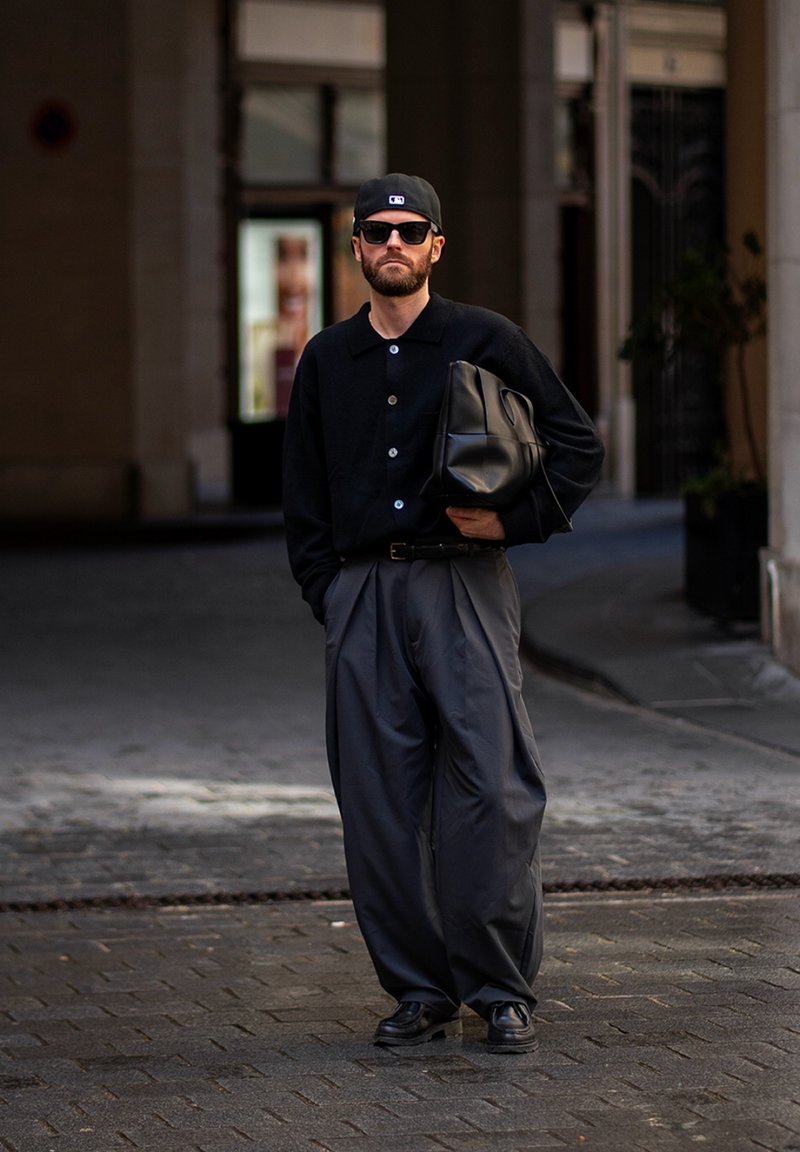 Homme portant une casquette noire, des lunettes de soleil, une chemise noire boutonnée, un pantalon large gris, tenant un sac noir, debout dans la rue de la ville.