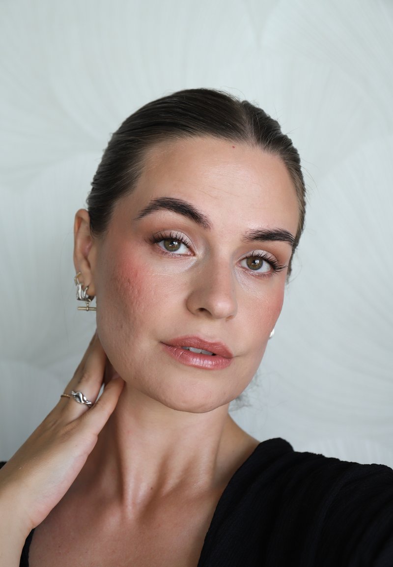 Mujer con ojos marrones y cabello oscuro recogido hacia atrás toca el cuello, lleva pendientes y anillos plateados, con maquillaje neutro y top negro.