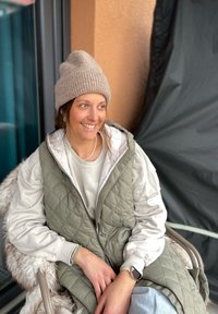 Κουρελιασμένο ελαιόχρωμο γιλέκο πάνω από ένα ανοιχτό γκρι φούτερ, συνδυασμένο με μια μπεζ πλεκτή σκούφo. Φοράει ένα ασημένιο βραχιόλι και ρολόι στον καρπό.