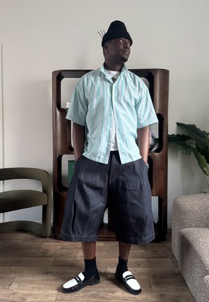 Hombre con camisa de manga corta a rayas, pantalones cortos anchos y oscuros hasta la rodilla y mocasines blanco y negro, de pie en el interior mirando hacia arriba a la derecha.