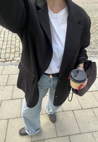 Personne portant un blazer noir, une chemise blanche, un jean bleu clair, tenant une tasse de café et un sac tressé, debout sur un trottoir pavé.