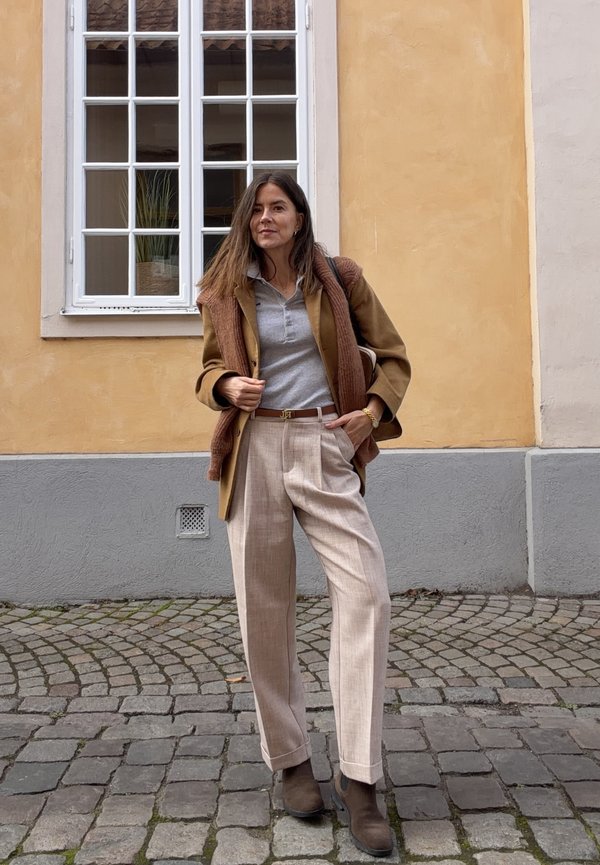 Mujer con pantalones beige, botas marrones, abrigo marrón y camisa gris, de pie sobre una calle adoquinada frente a una pared beige y gris con una ventana blanca.