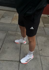 Białe buty sportowe z różowymi akcentami, czarnym logo i dzianinową cholewką, w połączeniu z czarnymi szortami z białym logo Adidas i białymi skarpetkami.