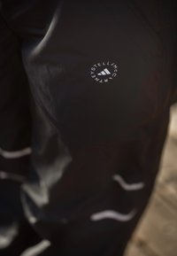 Imagem em close-up de calças de treino pretas com riscas brancas refletivas e um logótipo circular da Stella McCartney e Adidas na coxa.