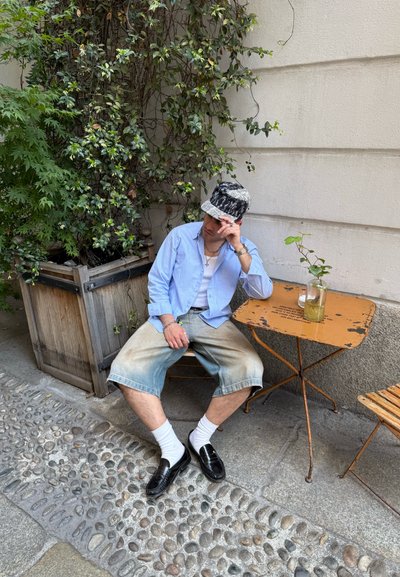 Hombre con sombrero tipo pescador, camisa azul claro y pantalones cortos vaqueros sentado en un banco junto a una pequeña mesa de metal con una planta en un frasco de cristal al aire libre.