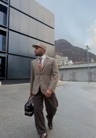 Hombre con blazer marrón, corbata estampada y gorra, llevando un bolso de cuero, caminando cerca de un edificio moderno con colinas al fondo.