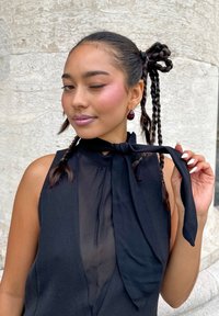 Vrouw met gevlochten haar dat in een strik is vastgebonden, gekleed in een mouwloze transparante zwarte top, knipogend met roze blush en glanzende lippenstift, terwijl ze aan het uiteinde van de strik houdt.