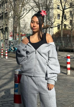 Joven con camiseta negra y chándal gris ajusta su chaqueta en una calle urbana soleada bordeada de árboles y bolardos.