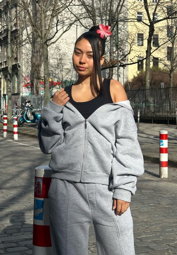 Jeune femme portant un haut noir et un survêtement gris ajuste sa veste dans une rue urbaine ensoleillée bordée d'arbres et de bornes.