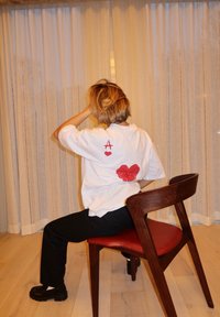 T-shirt bianco in cotone con grafica rossa di una "A" e un cuore sul retro. Indossato con pantaloni neri e scarpe nere voluminose. La sedia ha una struttura in legno.