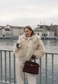 Veste en fausse fourrure beige, haut en tricot crème, pantalon blanc, gants en cuir marron, sac à main en daim bordeaux, tenant une tasse de café à motifs au bord de l'eau.