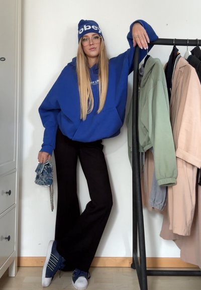 Sudadera azul de gran tamaño, pantalones anchos negros, gorro azul con el texto "obey", gafas transparentes, bolso de mezclilla, zapatillas blancas con rayas azules.